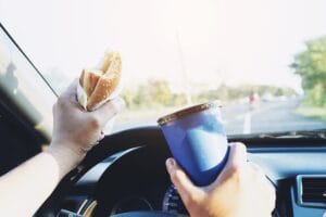 A man dangerously eats a hot dog and drinks a cold beverage while driving a car.